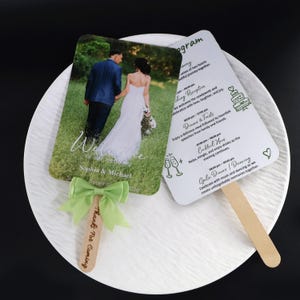 May include: Two wedding program fans with wooden handles. One fan features a photo of a couple in a blue suit and white dress on a grassy field with the text Welcome to our wedding Sophia & Michael. The other fan lists the event schedule.