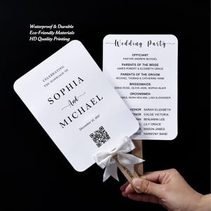May include: Two white rectangular wedding program fans with rounded corners, featuring black text and a QR code. The front displays "Celebrating the marriage of Sophia and Michael" with the date December 27, 2027. A white satin ribbon bow is tied to the handle.