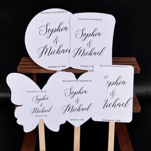 May include: Five white wedding hand fans with wooden handles, featuring black text that reads WELCOME TO THE WEDDING OF Sophia & Michael and the date December 27, 2026. The fans are shaped as a circle, a rectangle, a butterfly, and a heart.
