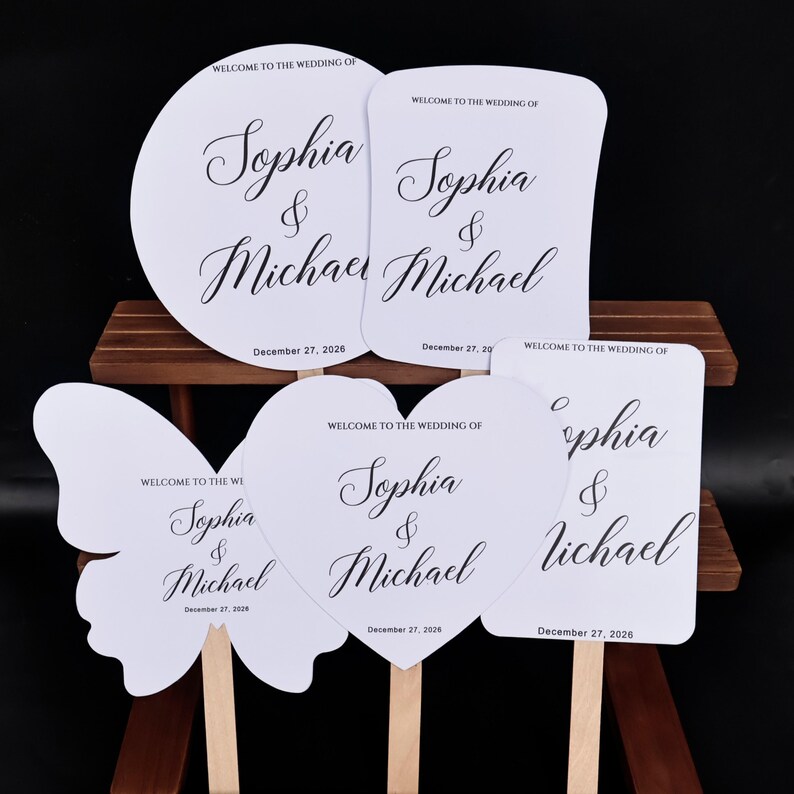May include: Five white wedding hand fans with wooden handles, featuring black text that reads WELCOME TO THE WEDDING OF Sophia & Michael and the date December 27, 2026. The fans are shaped as a circle, a rectangle, a butterfly, and a heart.