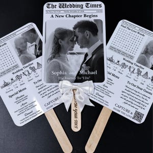 May include: Wedding-themed hand fans with a newspaper design. Each fan features a black and white photo of a couple, wedding details, and a menu. A white bow is attached to one fan, with custom text.