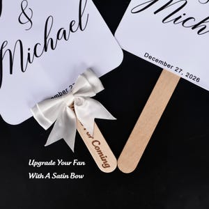 May include: Two white wedding fans with rounded corners, featuring black script text reading & Michael and the date December 27, 2026. One fan handle is plain wood, while the other features a cream satin bow and the text for Coming engraved.