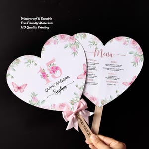 May include: Two heart-shaped paper fans featuring pink roses, green leaves, and butterflies. One fan displays the number 15, the text QUINCEAÑERA, and the name Sophia. The other fan shows a menu list. Both have light pink bows and wooden handles.