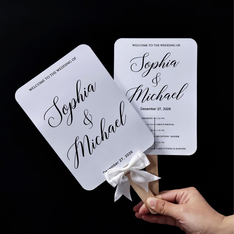 May include: Two white rectangular wedding program fans with rounded corners, featuring black text that reads Welcome to the Wedding of Sophia & Michael, December 27, 2026, and a schedule of events. A white satin ribbon bow is attached to the wooden handle.