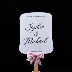 May include: A white rectangular hand fan with rounded corners featuring the black printed text WELCOME TO THE WEDDING OF, Sophia & Michael, and December 27, 2026. A light pink satin ribbon bow is tied around the wooden handle at the base.