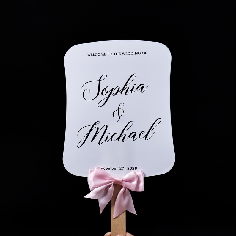 May include: A white rectangular hand fan with rounded corners featuring the black printed text WELCOME TO THE WEDDING OF, Sophia & Michael, and December 27, 2026. A light pink satin ribbon bow is tied around the wooden handle at the base.