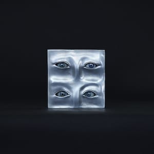 Puede incluir: Una escultura cuadrada de plata con cuatro ojos esculpidos en la superficie. Los ojos son azules y tienen un aspecto realista.