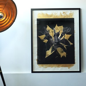 Puede incluir: Obra de arte enmarcada con fondo negro y detalles de pan de oro. El diseño abstracto presenta un rostro con elementos de papel rasgado. Una lámpara de estudio de estilo vintage con un gran reflector circular está en primer plano.