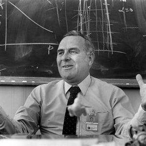 May include: A man with a white shirt and a dark tie is speaking with his hands raised. He is standing in front of a chalkboard with mathematical equations and diagrams written on it.