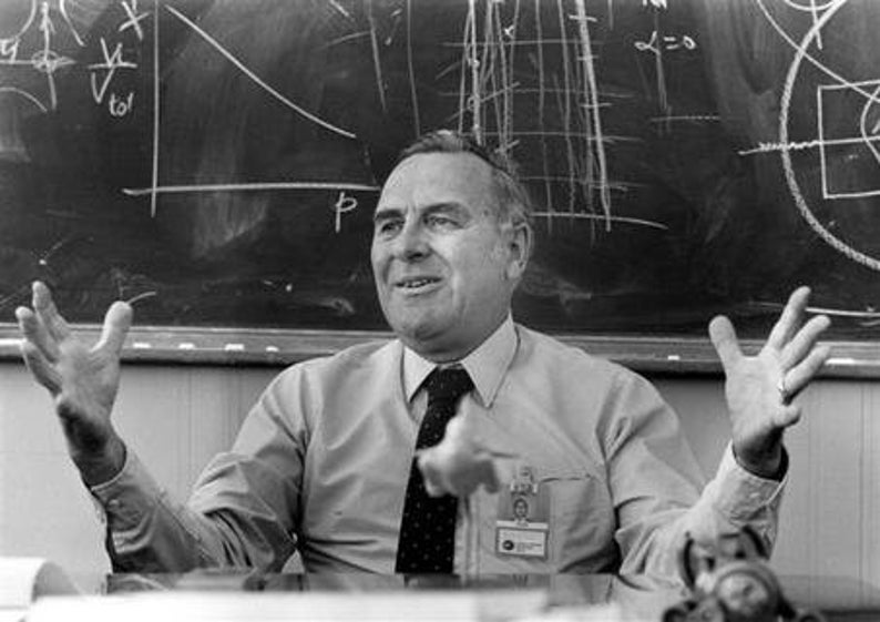 May include: A man with a white shirt and a dark tie is speaking with his hands raised. He is standing in front of a chalkboard with mathematical equations and diagrams written on it.
