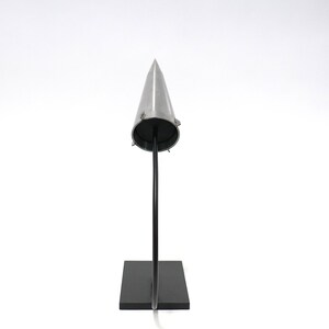 May include: A silver cone-shaped object on a black stand. The cone is pointed at the top and has a flat base. The stand is rectangular and has a curved metal rod that supports the cone.