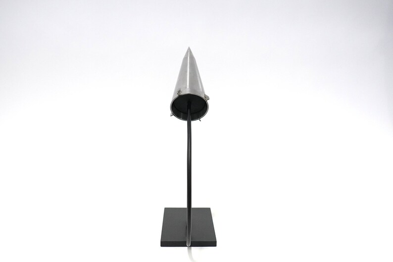 May include: A silver cone-shaped object on a black stand. The cone is pointed at the top and has a flat base. The stand is rectangular and has a curved metal rod that supports the cone.
