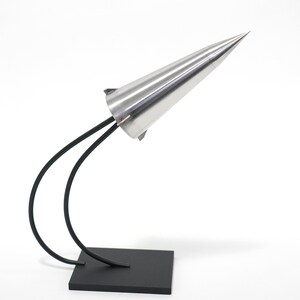 May include: A silver cone-shaped sculpture with a black metal base and two curved arms. The sculpture is on a white background.
