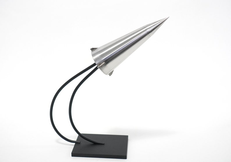 May include: A silver cone-shaped sculpture with a black metal base and two curved arms. The sculpture is on a white background.