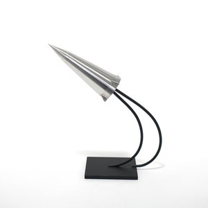 May include: A silver cone sculpture with a black metal base. The cone is mounted on two curved metal rods that are attached to the base.