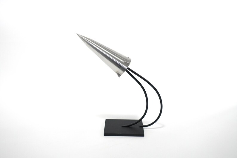 May include: A silver cone sculpture with a black metal base. The cone is mounted on two curved metal rods that are attached to the base.