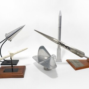 May include: Five model rockets on wooden bases. The rockets are made of metal and are painted silver. One rocket has the text "USAF" on the side.