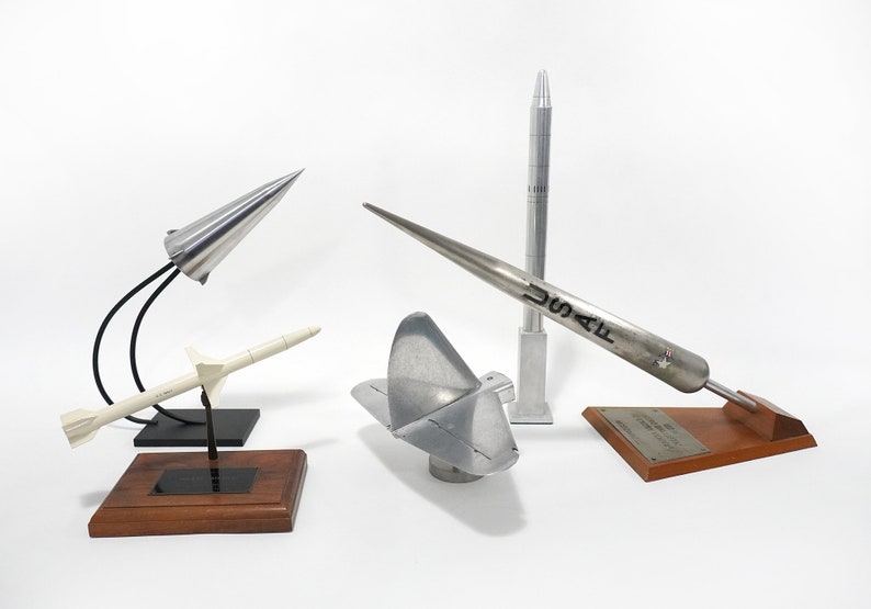 May include: Five model rockets on wooden bases. The rockets are made of metal and are painted silver. One rocket has the text "USAF" on the side.