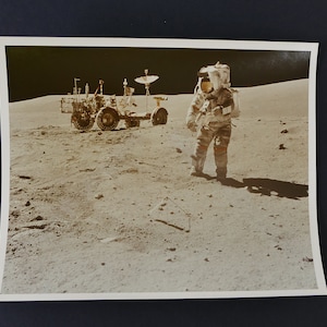 Puede incluir: Una fotografía en blanco y negro de un astronauta de pie en la superficie de la luna. El astronauta lleva un traje espacial blanco y un casco. El astronauta está de pie junto a un rover lunar, que es un pequeño vehículo de ruedas utilizado para explorar la superficie de la luna.