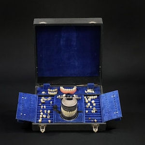 May include: A black wooden box with a blue velvet interior, containing a collection of dentures and dental tools. The box has a hinged lid and a metal latch.