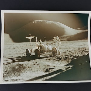 Puede incluir: Una fotografía en blanco y negro de un astronauta de pie junto a un rover lunar en la superficie de la luna. El rover tiene una gran antena parabólica en la parte superior. El astronauta lleva un traje espacial blanco y una mochila. La superficie de la luna está cubierta de cráteres y polvo.