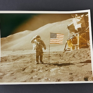 Puede incluir: Una fotografía en blanco y negro de un astronauta saludando la bandera estadounidense plantada en la superficie de la luna. El módulo lunar es visible en el fondo.