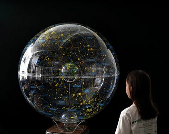 May include: A large, clear globe with a detailed map of the stars and constellations, illuminated with yellow and blue lines and dots. The globe sits on a clear stand. A person is looking at the globe.