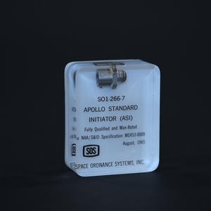 May include: A clear rectangular acrylic block containing a metallic component at the top. The text reads: SO1-266-7 APOLLO STANDARD INITIATOR (ASI) Fully Qualified and Man-Rated NAA/S&ID Specification MC453-0009 August, 1965 and SPACE ORDNANCE SYSTEMS, INC.