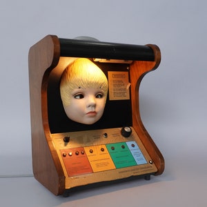 以下が含まれることがあります： 木製の箱の中に光があり、磁器製のドールヘッドを照らしています。箱にはノブとボタンがついたコントロールパネルと、「Chromatic Color Test Demonstration」と書かれたラベルが付いています。