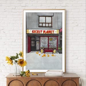 Puede incluir: Pintura a la acuarela de un escaparate con el texto "SECRET PLANET" en letras rojas. La tienda tiene una puerta roja y una ventana que muestra bicicletas. Un banco rojo está afuera. Un jarrón de girasoles y libros están en un mueble de madera.