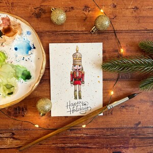 May include: A watercolor illustration of a nutcracker soldier on a textured card, with the words "Happy Holidays" in script. The image is surrounded by Christmas ornaments, a paintbrush, and a palette.