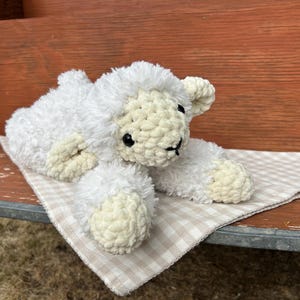 May include: A handmade, white crochet lamb plush toy with cream-colored accents, lying on a beige and white checkered cloth. The lamb has black eyes and a black stitched mouth. The plush is resting on a wooden bench.