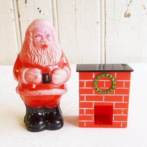 Pode incluir: Um conjunto de decoração de Natal vintage com uma figura vermelha do Papai Noel e uma lareira de tijolo vermelho com uma coroa verde. A figura do Papai Noel tem um rosto e barba rosa, botas pretas e um cinto preto.