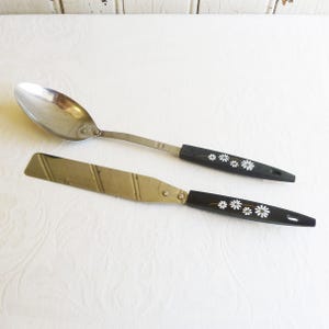 Puede incluir: Dos utensilios de cocina con mangos negros y patrones de margaritas blancas. Una cuchara de plata con un mango largo y una espátula de plata con un mango más corto.