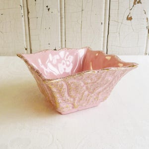 May include: A pink ceramic bowl with a square shape and scalloped edges. The bowl has a textured exterior with gold accents and a smooth, glossy interior. The rim is also trimmed with gold.