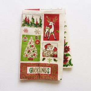 May include: A cream-colored fabric towel featuring a grid of Christmas-themed illustrations including red candles, a white reindeer, a decorated green Christmas tree, Santa Claus, and red poinsettias, with the word GREETINGS printed in green.