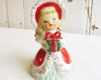 May include: A ceramic figurine of a person with blonde hair wearing a red bonnet with gold trim, a red and white coat with gold accents, and a light green skirt. The figure holds a green gift box with a red bow and has painted facial features.