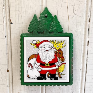 Può includere: Una placca decorativa da parete con cornice in metallo fuso verde e decorazione superiore a forma di pini. La piastrella centrale mostra un'illustrazione colorata di Babbo Natale in abito rosso con barba bianca, una renna, una slitta gialla e un sacco.