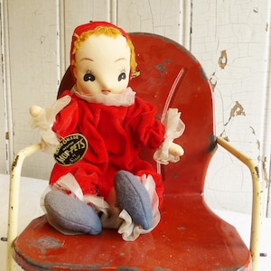 Puede incluir: Una muñeca vintage hecha a mano con un atuendo de terciopelo rojo, sentada en una silla de metal roja. La muñeca tiene una cara pintada, cabello rubio y zapatos azules. La muñeca tiene una etiqueta que dice "Handmade Mop-Pets by Serve".