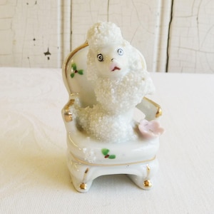 Puede incluir: Figura de caniche de porcelana blanca sentado en una silla a juego. El caniche tiene un pelaje texturizado y esponjoso, boca rosa y ojos negros. La silla está decorada con ribetes dorados y pequeños detalles florales verdes y rosas.