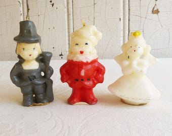 May include: Three vintage figurine candles. One is a pilgrim in a gray outfit and hat, the second is a Santa Claus figure in a red suit, and the third is a princess in a white dress. The candles are on a white surface.