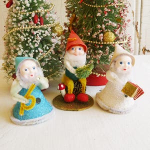 Puede incluir: Tres figuras de gnomos navideños vintage. Un gnomo lleva un gorro rojo y un chaleco verde, sosteniendo un arbolito. Otro gnomo lleva un vestido azul y sostiene un instrumento amarillo. El tercer gnomo lleva un vestido blanco y sostiene un libro dorado.