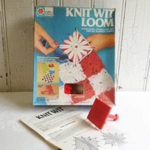May include: A vintage Knit Wit Loom craft kit in a blue cardboard box featuring red and white yarn flower motifs. The set includes a red plastic square loom tool with metal pins and a printed instruction manual with diagrams for yarn projects.