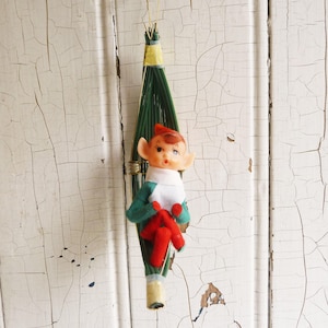 May include: A vintage Christmas ornament featuring a green and white elf doll sitting in a green straw basket. The elf has a red hat, green and white clothing, and red shoes.