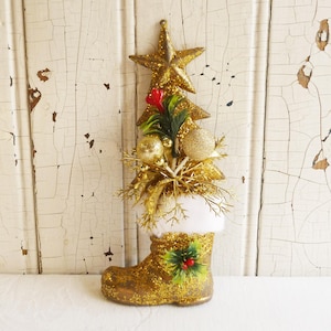 May include: A gold glitter Christmas boot ornament topped with a star, tree, and festive decorations. The boot has a white faux fur trim and is adorned with a holly sprig. The ornament is designed for holiday decor.