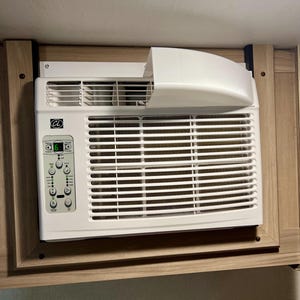 May include: A white air conditioning unit installed in a wooden cabinet. The unit has a control panel with buttons and a digital display. The top of the unit has a white cover. The unit is mounted on a wall.