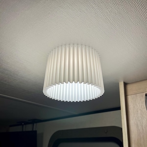 May include: A white, pleated lampshade hangs from the ceiling. The shade has a cylindrical shape with vertical pleats and a scalloped bottom edge. The light fixture is mounted on a white ceiling.