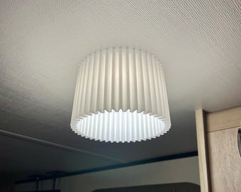 May include: A white, pleated lampshade hangs from the ceiling. The shade has a cylindrical shape with vertical pleats and a scalloped bottom edge. The light fixture is mounted on a white ceiling.