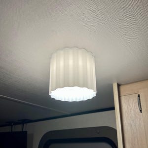 May include: A white, cylindrical light fixture with a scalloped edge is mounted on a textured ceiling. The light has vertical ridges and emits a soft glow. The fixture is in a room with wood-toned cabinetry.
