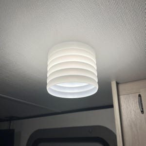 May include: A white, cylindrical light fixture with a layered design is mounted on a white ceiling. The fixture emits a soft, diffused light, creating a warm ambiance. The design features multiple horizontal, circular bands.